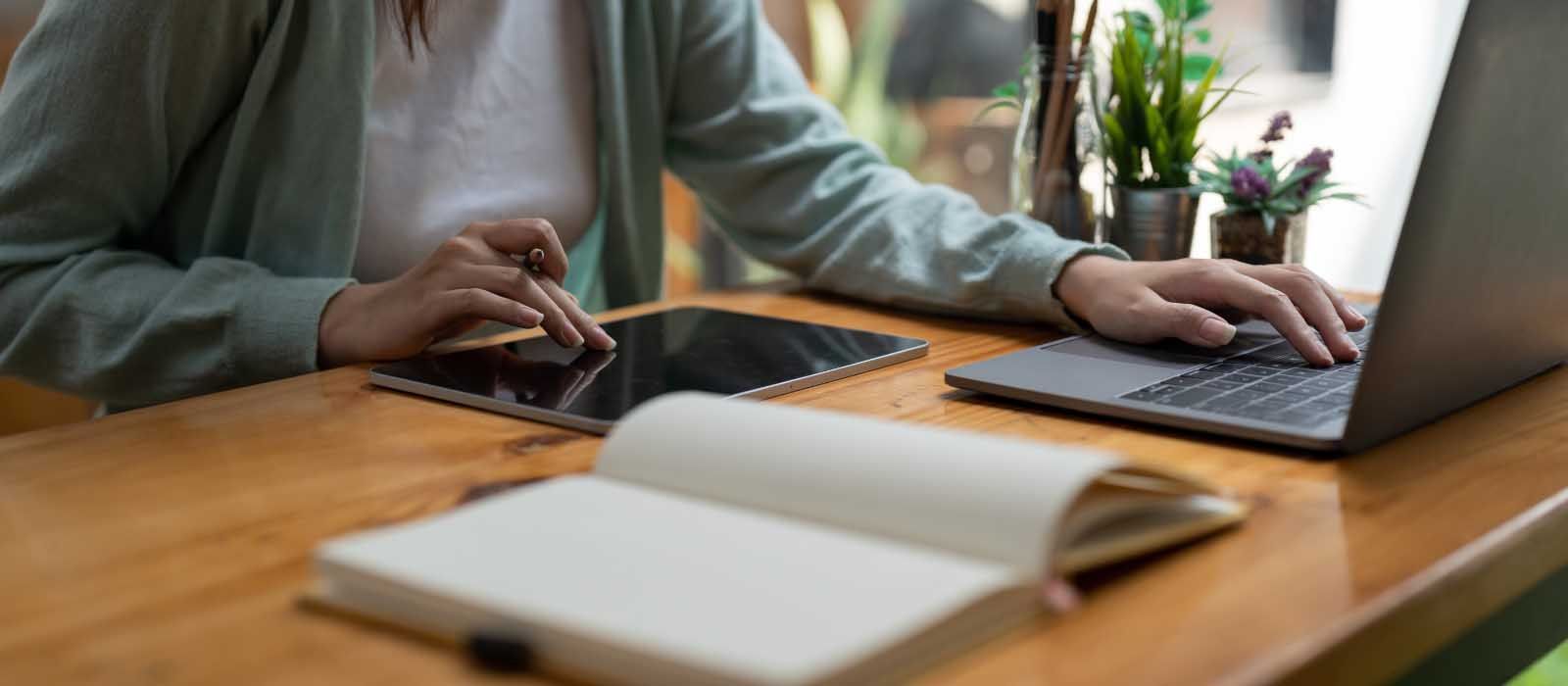 A woman works on her laptop and tablet, seated at a wooden table, focused on her tasks.